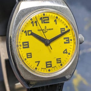 May include: A vintage Ulysse Nardin wristwatch with a bright yellow face and black hands and numerals. The watch has a silver-colored case and a black leather band. The watch face displays the brand name and the word "SWISS MADE".