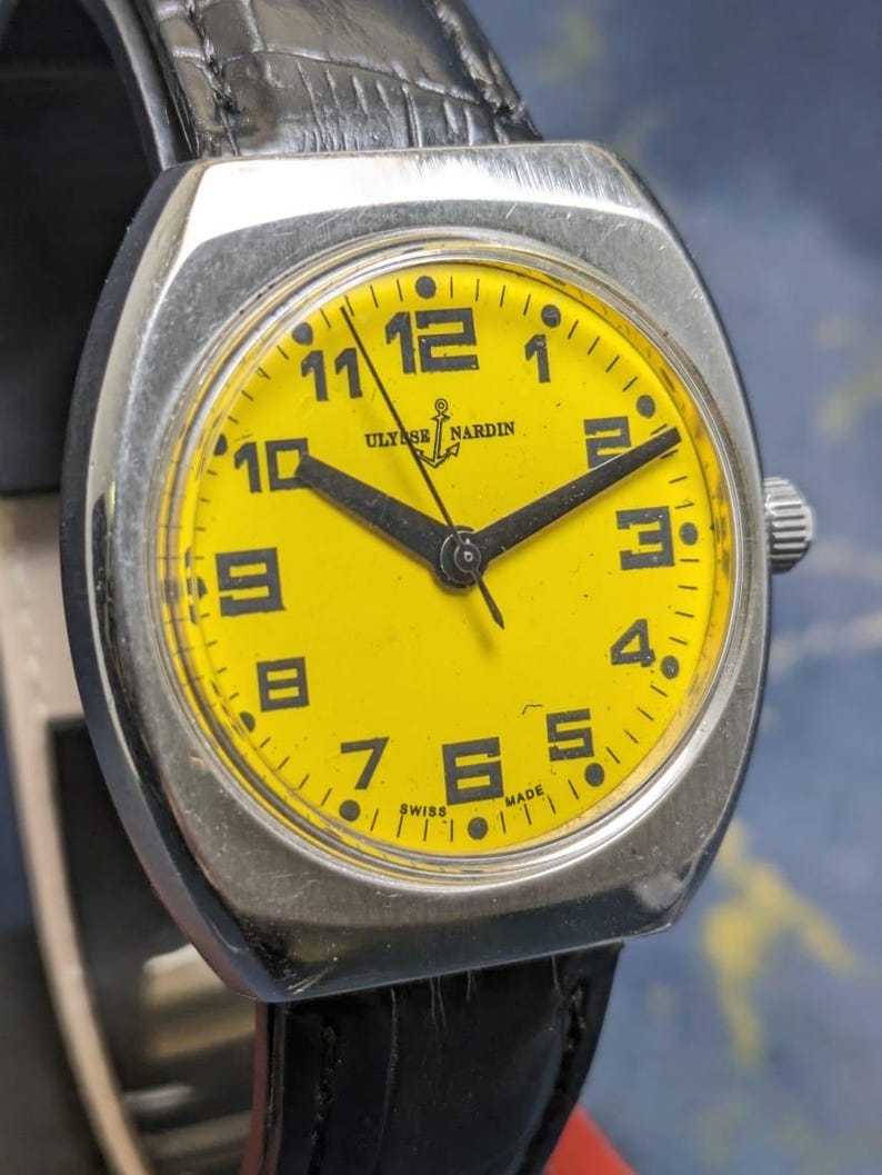 May include: A vintage Ulysse Nardin wristwatch with a bright yellow face and black hands and numerals. The watch has a silver-colored case and a black leather band. The watch face displays the brand name and the word "SWISS MADE".