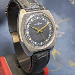 May include: Vintage Ulysse Nardin watch with a black dial, gold hands, and a stainless steel case. The watch features a black leather band and is marked "Swiss Made". The watch has a rectangular face.