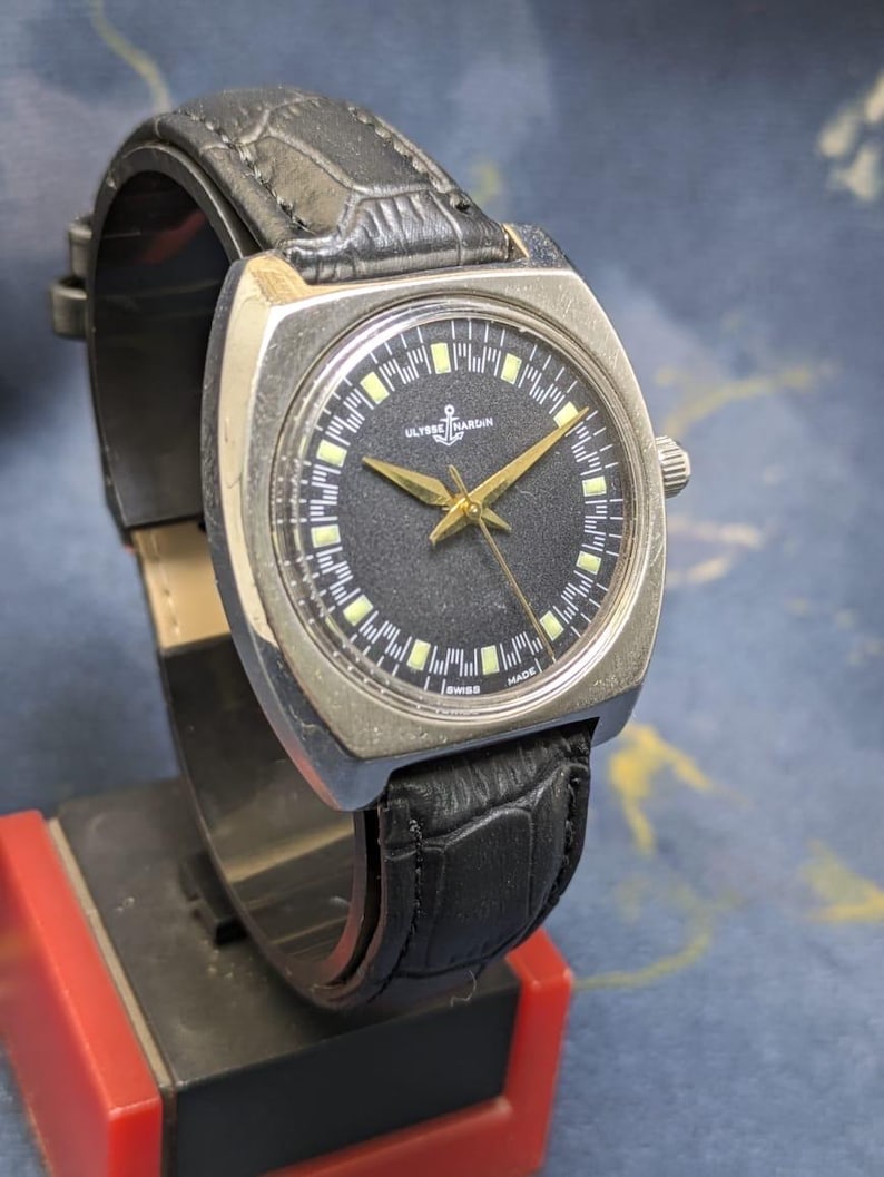 May include: Vintage Ulysse Nardin watch with a black dial, gold hands, and a stainless steel case. The watch features a black leather band and is marked "Swiss Made". The watch has a rectangular face.