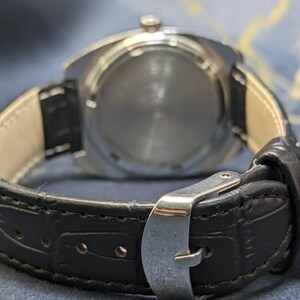 May include: Close-up of a wristwatch, showcasing the back of the watch and the black leather band. The watch has a silver-colored case and a black leather strap with a silver buckle. The band is approximately 0.75 inches wide.