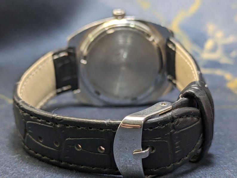 May include: Close-up of a wristwatch, showcasing the back of the watch and the black leather band. The watch has a silver-colored case and a black leather strap with a silver buckle. The band is approximately 0.75 inches wide.