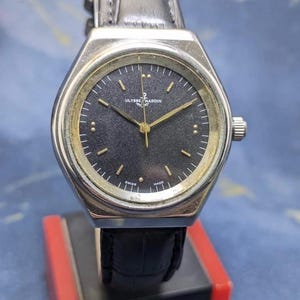 May include: A silver Ulysse Nardin watch with a black face and gold hands. The watch has a leather strap and is displayed on a red and black stand. The watch face has small gold markers and the words "SWISS MADE".