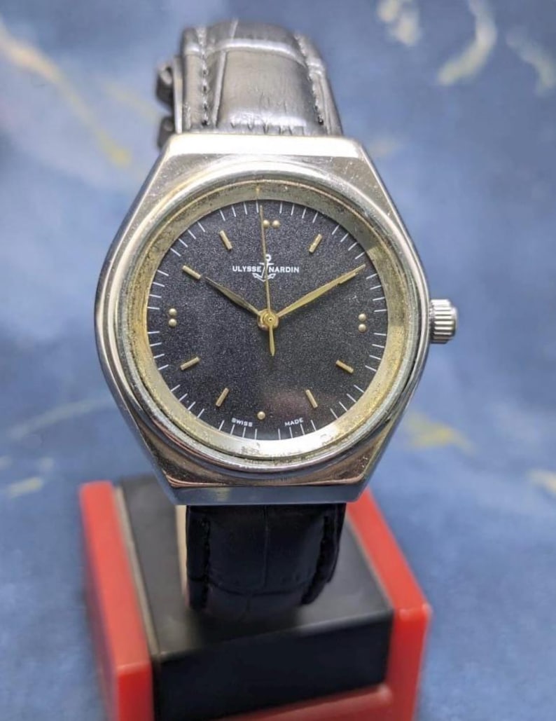May include: A silver-toned Ulysse Nardin watch with a black face and gold hands. The watch has a leather band and is displayed on a red and black stand. The watch face has small gold markers and the words "SWISS MADE".