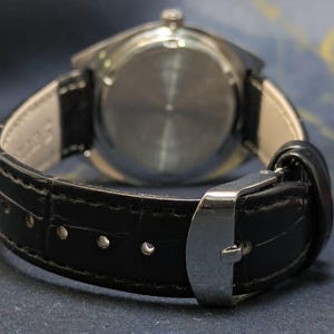 May include: Close-up of a wristwatch with a black leather band and a silver clasp. The watch face is not visible. The band is stitched with white thread. The watch is a classic accessory.