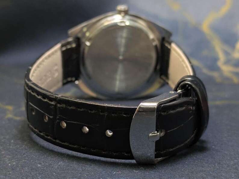 May include: Close-up of a wristwatch with a black leather band and a silver clasp. The watch face is not visible. The band is stitched with white thread. The watch is a classic accessory.