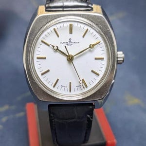 May include: A vintage Ulysse Nardin wristwatch with a silver-tone case and a white dial. The watch features gold-tone hands and markers, a black leather strap, and a crown on the right side. The watch is displayed on a red and black stand.