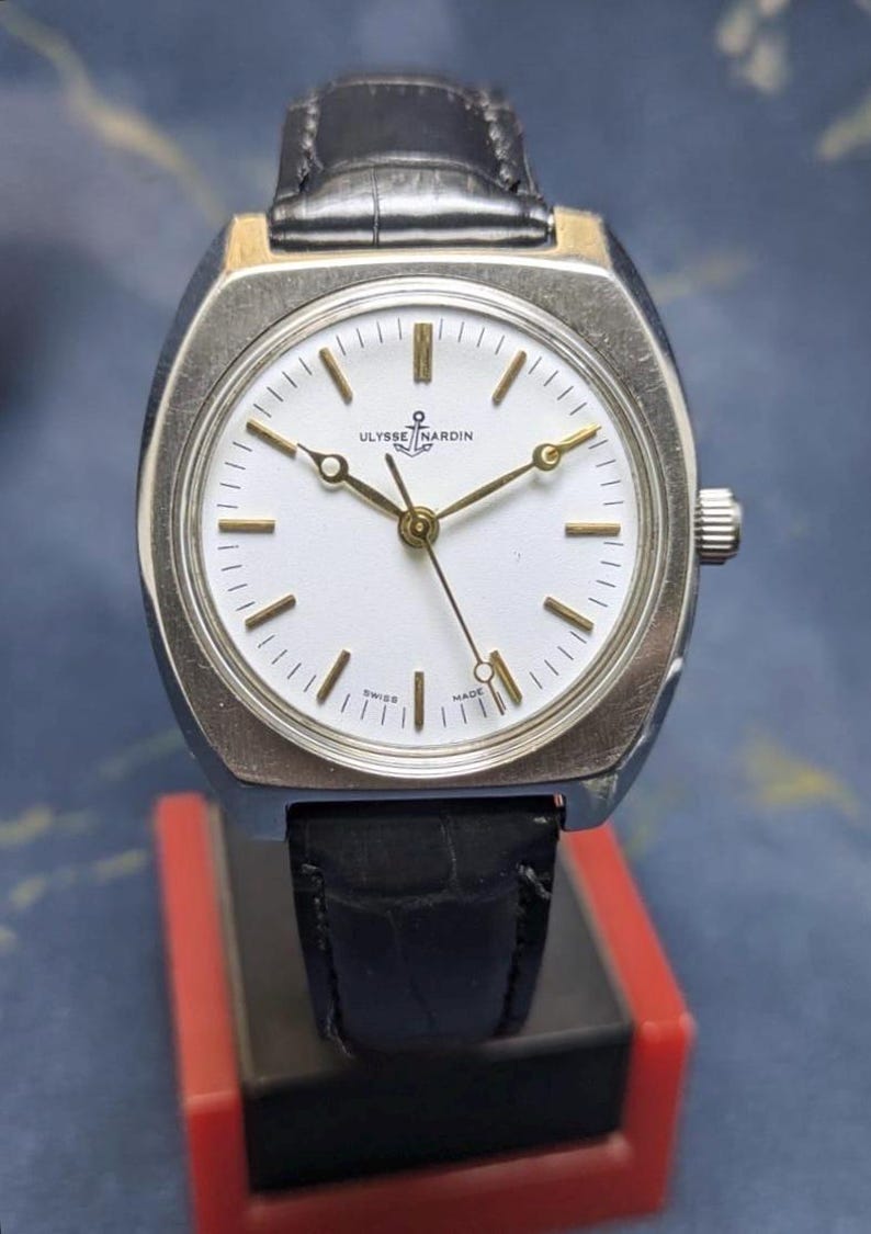May include: A vintage Ulysse Nardin wristwatch with a silver-tone case and a white dial. The watch features gold-tone hands and markers, a black leather strap, and a crown on the right side. The watch is displayed on a red and black stand.