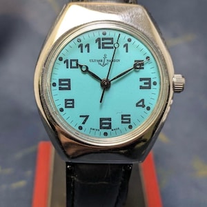 May include: A vintage Ulysse Nardin watch with a light blue face and black hands. The watch has a silver-colored case and a black leather band. The watch face displays the numbers 1 through 12, with the brand name and "SWISS MADE" visible.