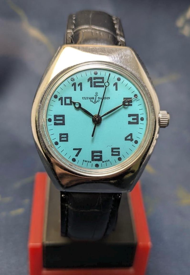 May include: A vintage Ulysse Nardin watch with a light blue face and black hands. The watch has a silver-colored case and a black leather band. The watch face displays the numbers 1 through 12, with the brand name and "SWISS MADE" visible.
