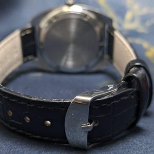 May include: Close-up of a wristwatch with a black leather band and a silver buckle. The watch face is visible in the background. The band has a textured pattern.