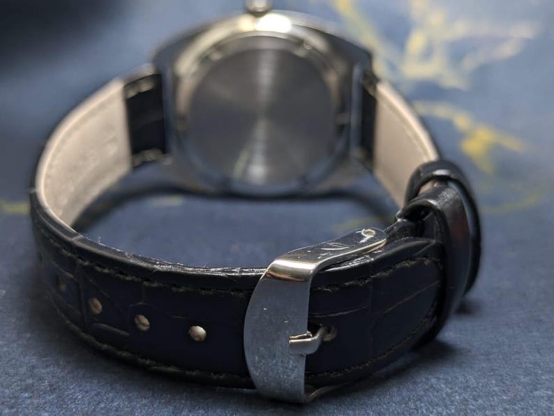 May include: Close-up of a wristwatch with a black leather band and a silver buckle. The watch face is visible in the background. The band has a textured pattern.