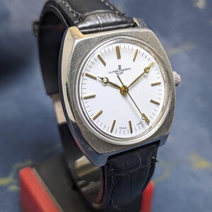 May include: A vintage Ulysse Nardin watch with a white face, gold hands, and a black leather band. The watch has a silver-colored square case and is displayed on a red and black stand. The watch face has the text "Ulysse Nardin" and "Swiss".