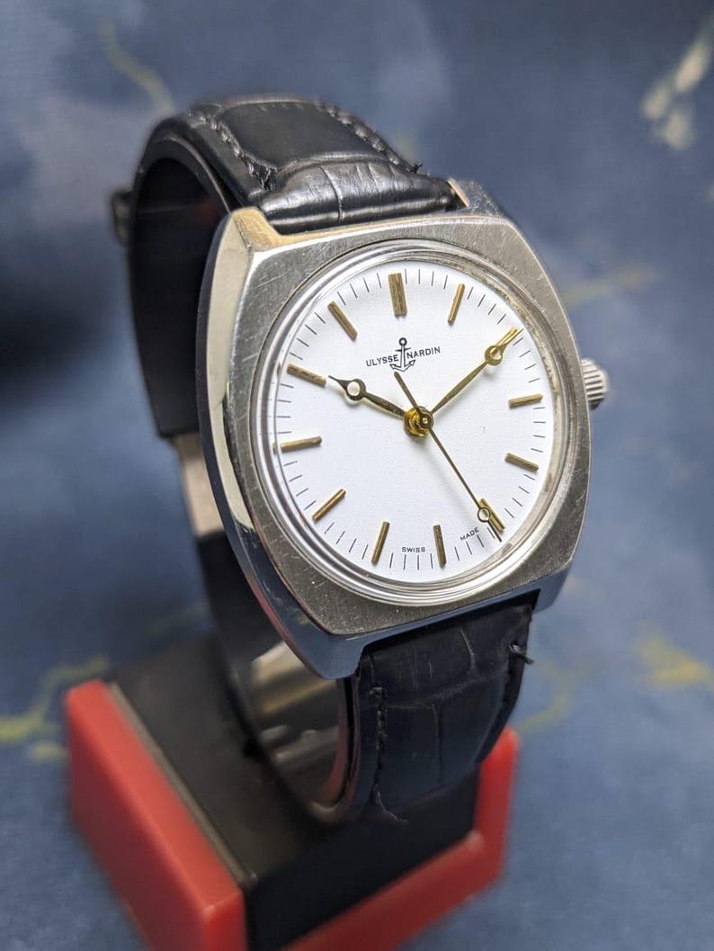 May include: A vintage Ulysse Nardin watch with a white face, gold hands, and a black leather band. The watch has a silver-colored square case and is displayed on a red and black stand. The watch face has the text "Ulysse Nardin" and "Swiss".