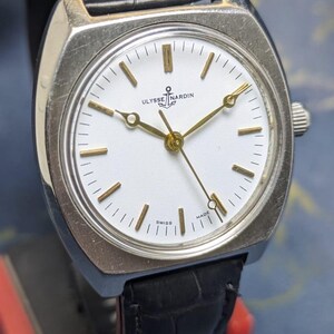 May include: A vintage Ulysse Nardin wristwatch with a silver-toned case and a white face. The watch features gold-colored hands and markers, and a black leather band. The watch face displays the brand name and "Swiss Made".
