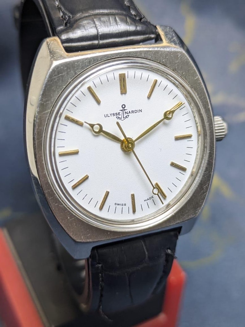 May include: A vintage Ulysse Nardin wristwatch with a silver-toned case and a white face. The watch features gold-colored hands and markers, and a black leather band. The watch face displays the brand name and "Swiss Made".