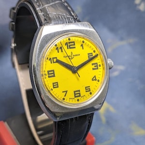 May include: A vintage Ulysse Nardin wristwatch with a bright yellow face and black numerals. The watch has a silver-toned case and a black leather band. The watch is displayed on a black stand.