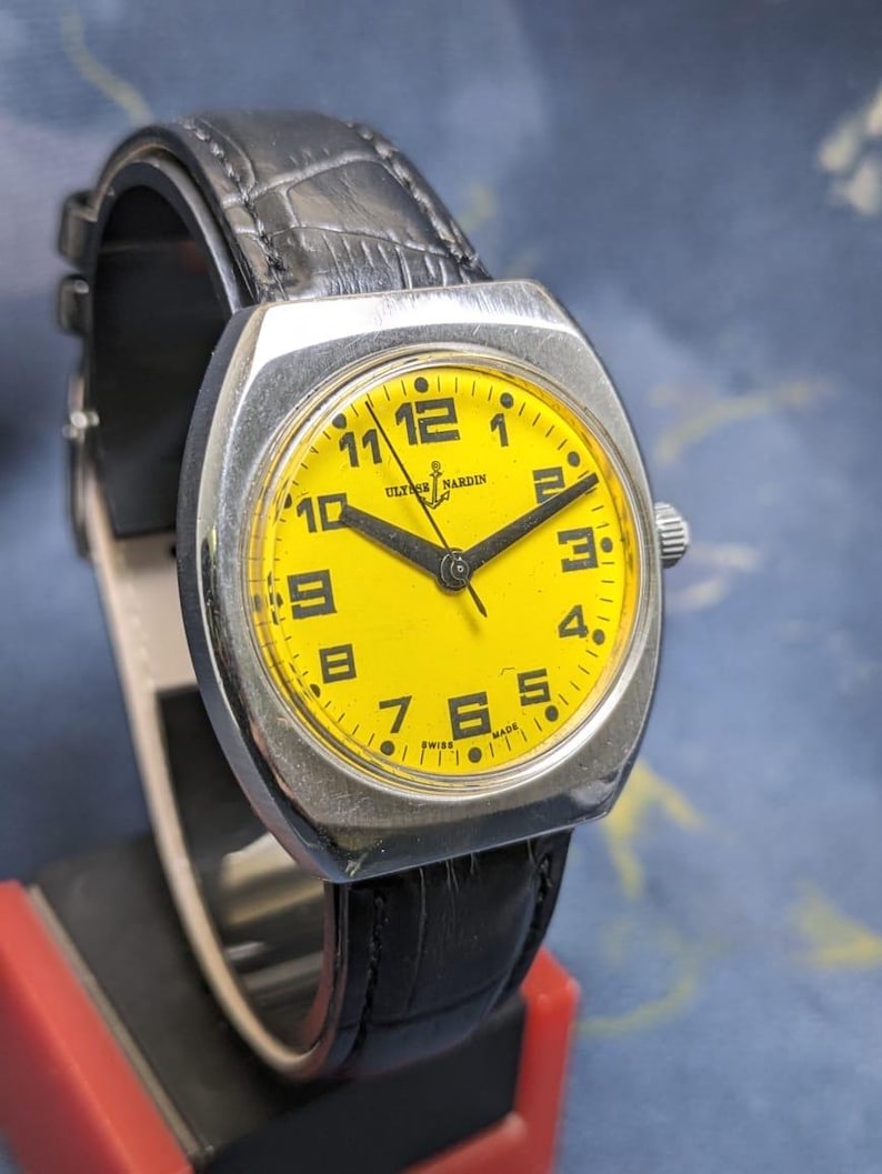 May include: A vintage Ulysse Nardin wristwatch with a bright yellow face and black numerals. The watch has a silver-toned case and a black leather band. The watch is displayed on a black stand.