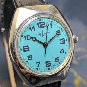 May include: A silver-toned Ulysse Nardin wristwatch with a turquoise face and black numerals. The watch has a black leather band and the words "ULYSSE NARDIN" and "SWISS MADE" are visible on the watch face.