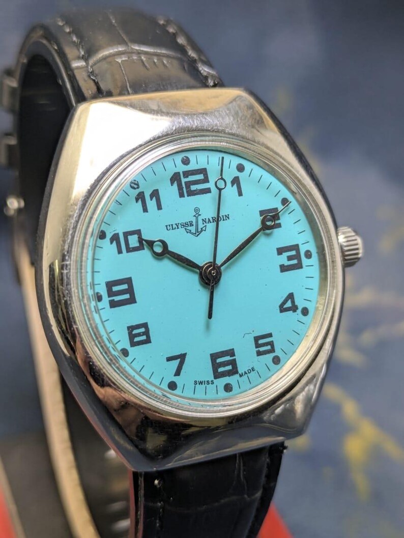 May include: A silver-toned Ulysse Nardin wristwatch with a turquoise face and black numerals. The watch has a black leather band and the words "ULYSSE NARDIN" and "SWISS MADE" are visible on the watch face.