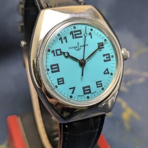 May include: A vintage Ulysse Nardin watch with a turquoise face, black numerals, and black hands. The watch has a silver-colored case and a black leather band. The watch face displays the brand name and the words "SWISS MADE".