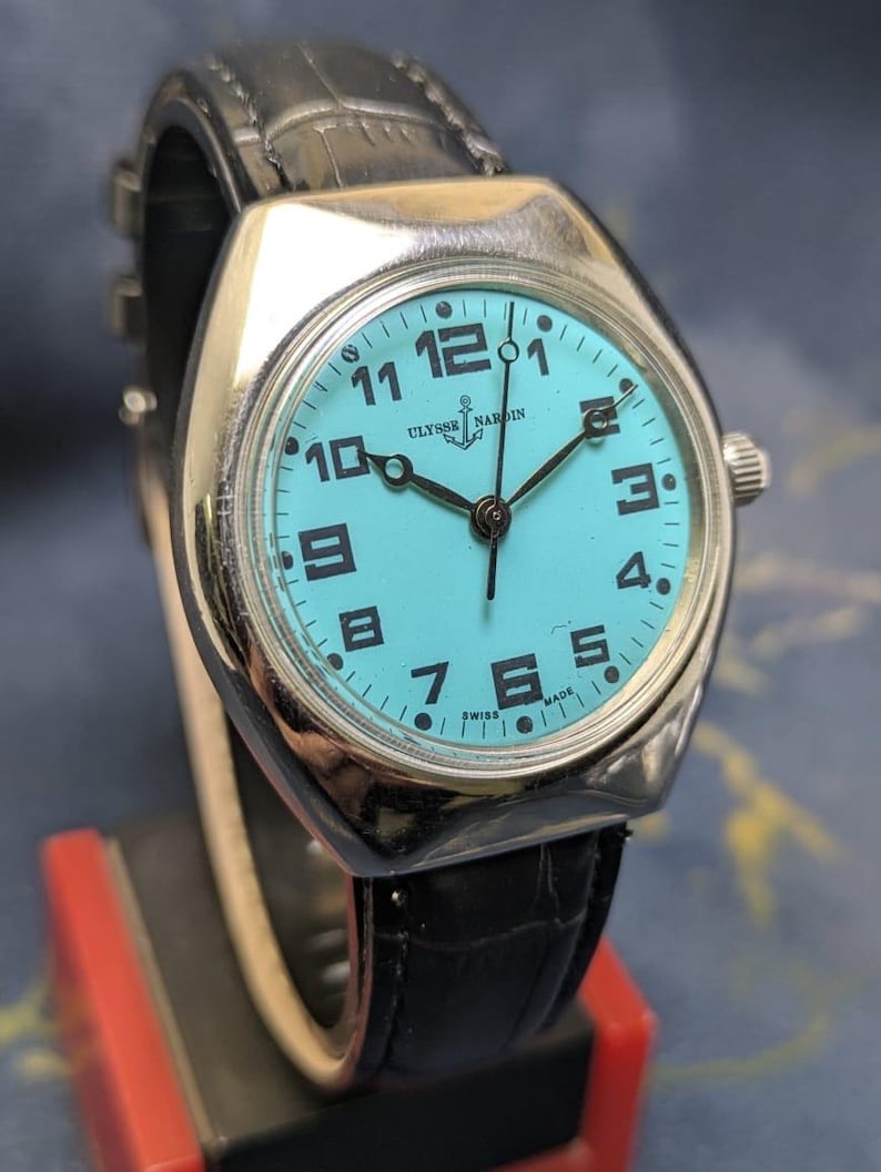 May include: A vintage Ulysse Nardin watch with a turquoise face, black numerals, and black hands. The watch has a silver-colored case and a black leather band. The watch face displays the brand name and the words "SWISS MADE".