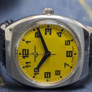 May include: A vintage Ulysse Nardin wristwatch with a bright yellow face and black numerals. The watch has a silver-toned case and black hands. The watch is attached to a black leather band. The watch face reads "Ulysse Nardin" and "Swiss Made".