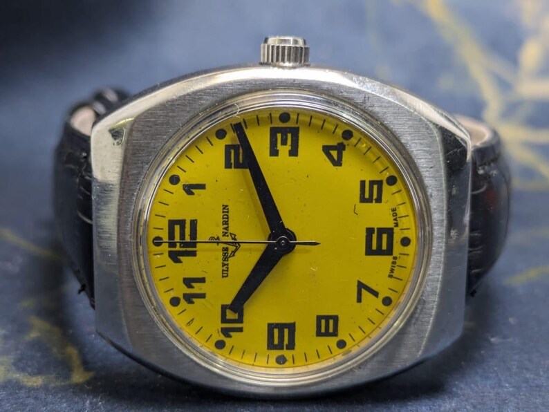 May include: A vintage Ulysse Nardin wristwatch with a bright yellow face and black numerals. The watch has a silver-toned case and black hands. The watch is attached to a black leather band. The watch face reads "Ulysse Nardin" and "Swiss Made".