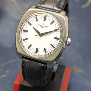 May include: A silver-toned wristwatch with a white face and black hands and markers. The watch features the brand name "Ulysse Nardin" and a small anchor logo. It has a black leather band and a silver crown.