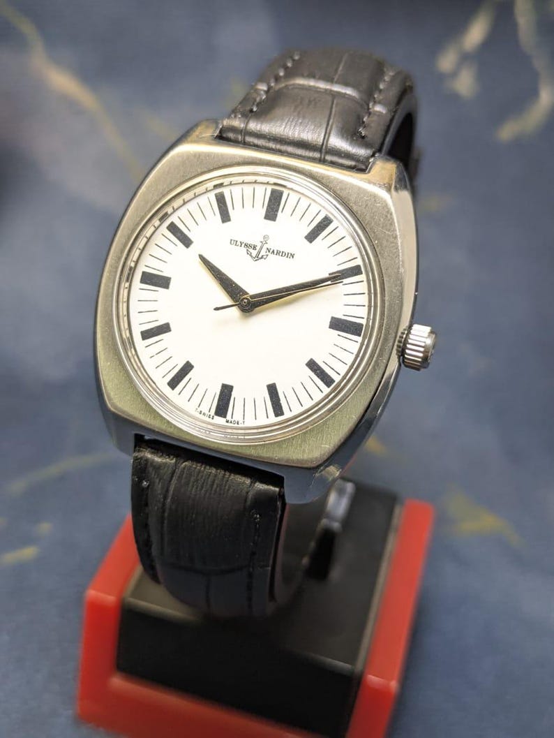 May include: A silver-toned wristwatch with a white face and black hands and markers. The watch features the brand name "Ulysse Nardin" and a small anchor logo. It has a black leather band and a silver crown.