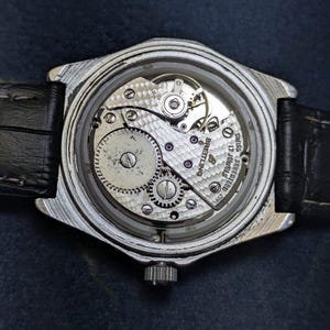 May include: A silver-toned wristwatch with a black leather band. The watch's back is open, revealing the intricate mechanical movement with gears, screws, and the Breitling logo. The watch face is not visible.
