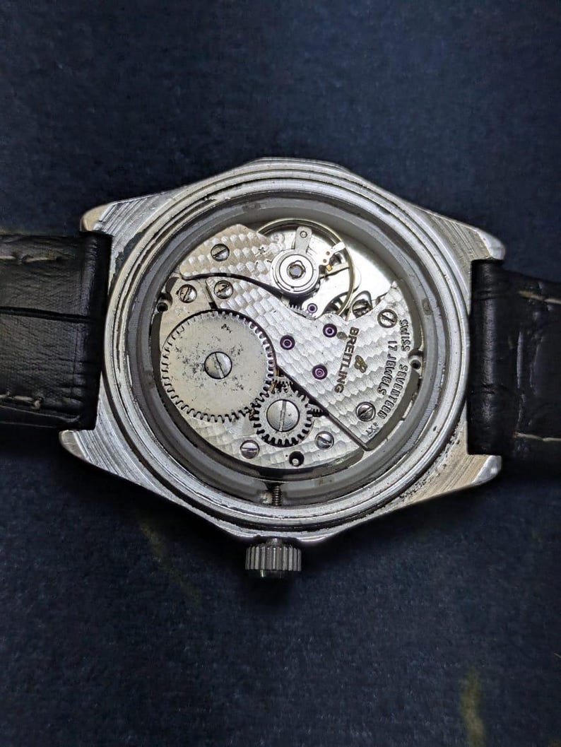 May include: A silver-toned wristwatch with a black leather band. The watch's back is open, revealing the intricate mechanical movement with gears, screws, and the Breitling logo. The watch face is not visible.