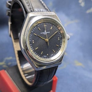 May include: A vintage Ulysse Nardin watch with a black dial and gold hands. The watch has a silver-tone case and a black leather strap. The watch is displayed on a red and black stand.