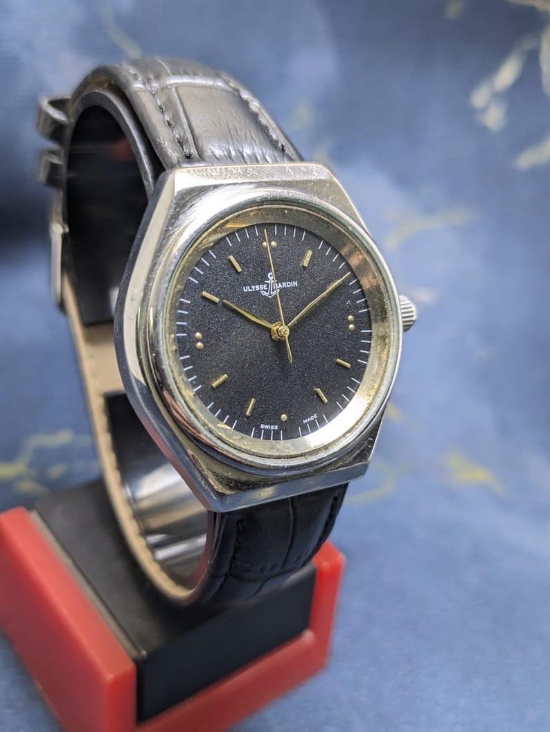 May include: A vintage Ulysse Nardin watch with a black dial and gold hands. The watch has a silver-tone case and a black leather strap. The watch is displayed on a red and black stand.