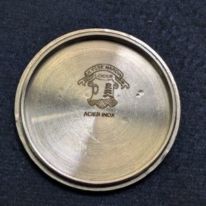 May include: A circular, brushed metal watch back with the engraved text "ULYSSE NARDIN LOCLE" above a crest design. The text "ACIER INOX" is engraved below the crest. The watch back is a light gold color.