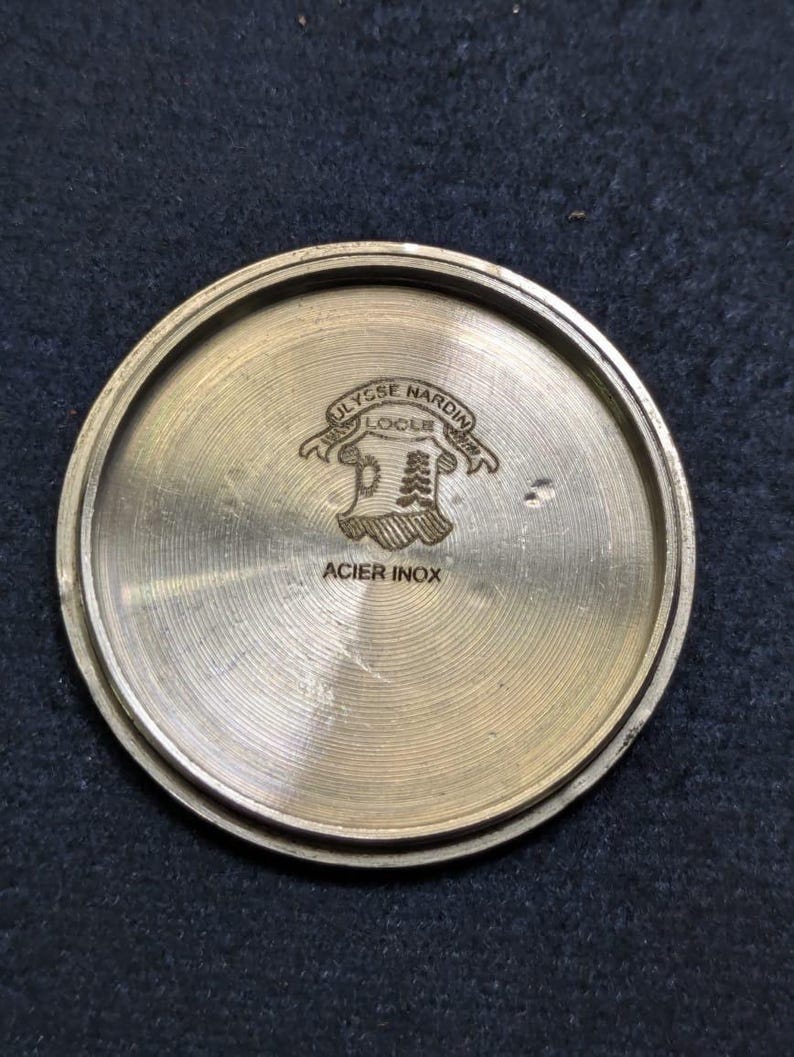 May include: A circular, brushed metal watch back with the engraved text "ULYSSE NARDIN LOCLE" above a crest design. The text "ACIER INOX" is engraved below the crest. The watch back is a light gold color.