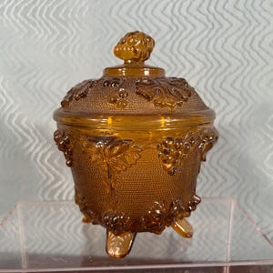 May include: Amber-colored glass jar with a lid, featuring a textured surface and grape cluster designs. The jar has three small feet and a decorative knob on the lid. A vintage-style decorative container.