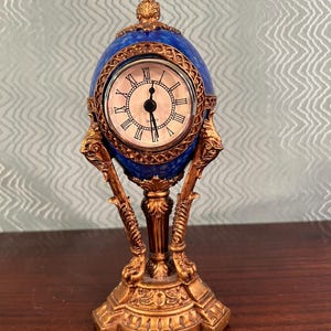 May include: A table clock shaped like an egg with a blue mottled finish and gold-colored metal accents. The clock face features a white background with black Roman numerals and black hands. The base and frame are decorated with ornate gold-colored metal.