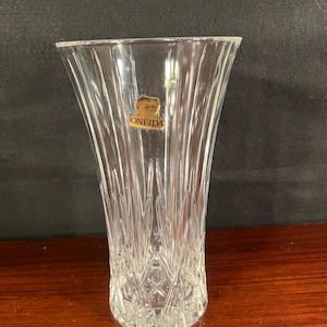 May include: A clear glass vase featuring a flared rim and a tapered body with vertical and crisscross cut patterns. A gold-colored sticker is attached to the upper portion of the glass, displaying the text Capri and the brand name ONEIDA.
