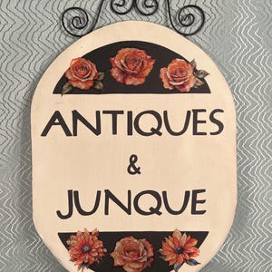May include: Oval wooden sign with the words "ANTIQUES & JUNQUE" in black lettering. The sign features three orange roses at the top and three floral designs at the bottom, set against a black background. A decorative black metal scroll is at the top.