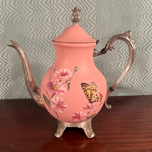 May include: A pink teapot with a silver handle, spout, and lid. The teapot is decorated with pink flowers and a butterfly. The teapot sits on three silver feet. A decorative item for tea parties.