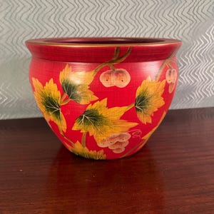 May include: A round, red planter featuring a painted pattern of yellow and green grape leaves and clusters of grapes. The bowl-shaped vessel has a smooth, dark red interior rim and a textured exterior surface with a visible wood grain finish.