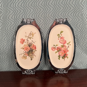 May include: Two vintage oval trays with silver-colored ornate frames. Each tray features a cream-colored center with a floral design of pink and peach roses and green leaves. The trays are displayed on a dark brown surface.
