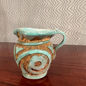 May include: A small, handcrafted ceramic pitcher with a turquoise glaze and brown accents. The pitcher features a handle and decorative carved patterns around the body. The interior is a lighter turquoise color.