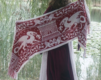 May include: A woman wearing a long, burgundy dress with a white knitted shawl featuring a red and white dragon design. The shawl has a white castle and other intricate details.