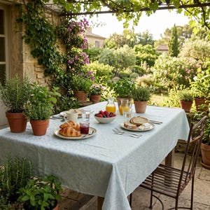 Puede incluir: Una mesa preparada para una comida al aire libre, cubierta con un mantel azul claro estampado. La mesa está puesta con platos de comida, vasos y un cuenco de bayas. Hierbas y plantas en macetas rodean la mesa, con un exuberante jardín al fondo.