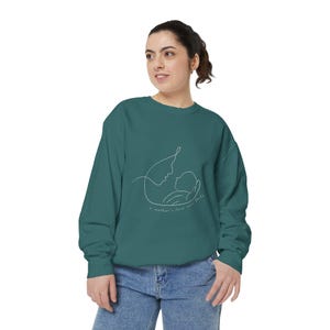 Könnte beinhalten: Ein blaugrünes Langarm-Sweatshirt mit einer weißen Strichzeichnung, die einen Elternteil zeigt, der ein Baby wiegt. Unter der Illustration steht der Text "a mother's love never fails" in einer zarten Schreibschrift. Rundhalsausschnitt.
