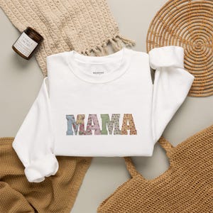 May include: A white crewneck sweatshirt featuring the word MAMA printed in a block font. Each letter is filled with a different colorful floral and gingham pattern. The sweatshirt has long sleeves and a ribbed collar, cuffs, and hemline.