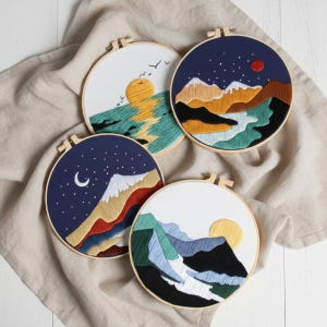 Op de afbeelding: Vier ronde borduurringen met landschapsontwerpen. Eén toont een zonsondergang boven water met vogels, een ander een bergketen onder een sterrenhemel, een derde een berglandschap met een halve maan en de laatste een berglandschap met een ondergaande zon.