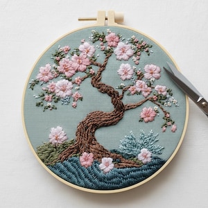 Könnte beinhalten: Handgestickte Stickerei eines blühenden Baumes in einem runden Holzrahmen. Der Baum hat einen braunen Stamm und Äste mit rosa und weißen Blüten. Der Hintergrund ist ein hellblaugrüner Stoff. Eine Schere befindet sich auf der rechten Seite.